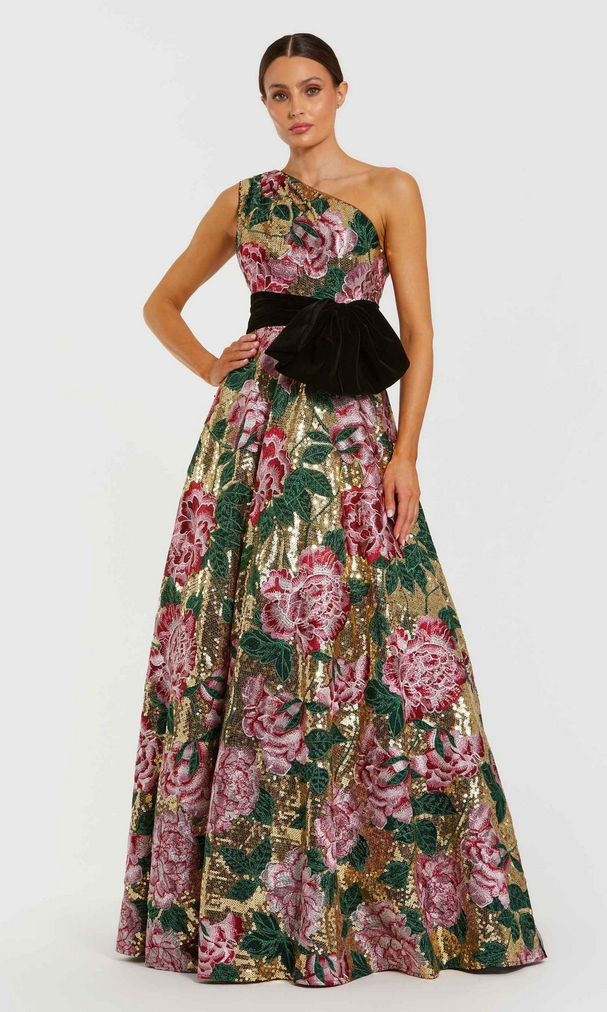 A woman wears the Mac Duggal 21194 long formal dress in Gold Multi, a one-shoulder, floor-length sequin ballgown with a floral-embroidered pattern and a large black waist bow.